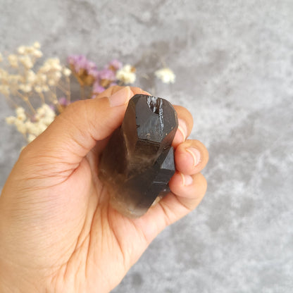 A hand holding a dark brown smoky quartz crystal point against a textured gray background with blurred dried flowers.