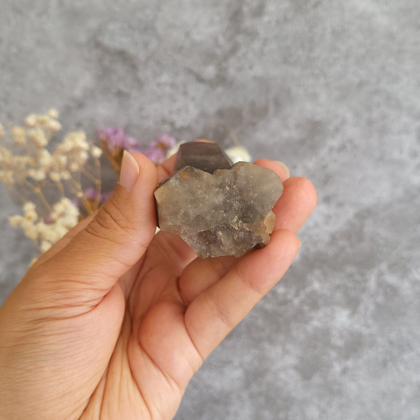 A hand holding a raw, rough gray crystal cluster against a textured gray background with blurred dried flowers.