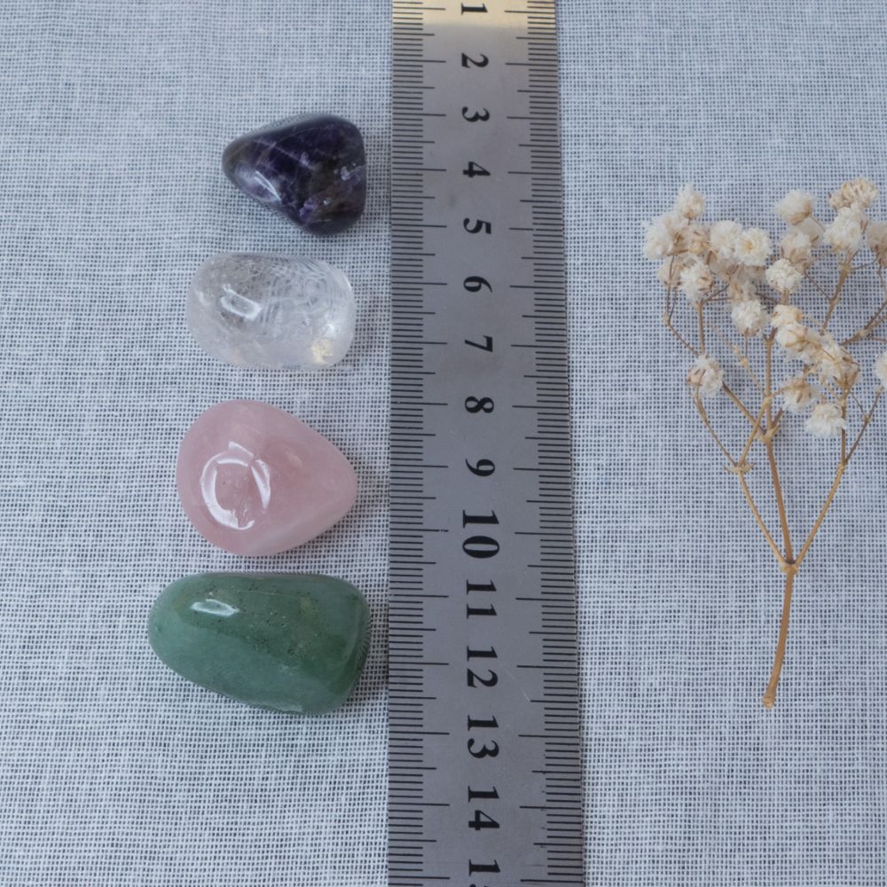 Four crystals of different colors (purple, clear, pink, green) next to a ruler on a textured fabric background.