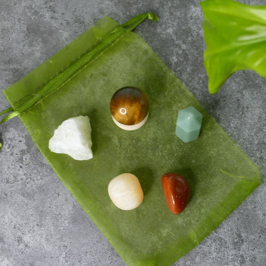 Set of raw lemon quartz, tiger's eye sphere, green aventurine mini tower, orange calcite and red jasper tumbled stones on a green drawstring bag against a gray concrete background