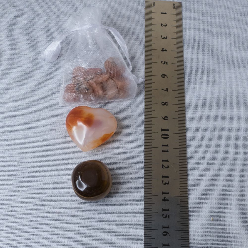 a sunstone chip pouch, a carnelian heart and a tiger's eye tumble stone on a textured surface with a ruler for scale.