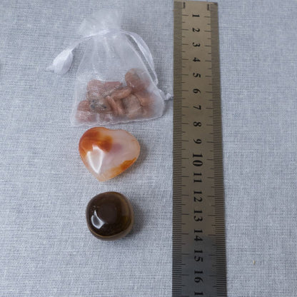 a sunstone chip pouch, a carnelian heart and a tiger's eye tumble stone on a textured surface with a ruler for scale.