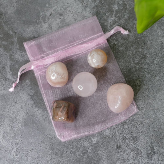 Set of pink amethyst, rhodonite, rose quartz tumbled stones, flower agate mushroom and rose quartz mini sphere on a pink drawstring bag on a gray surface 