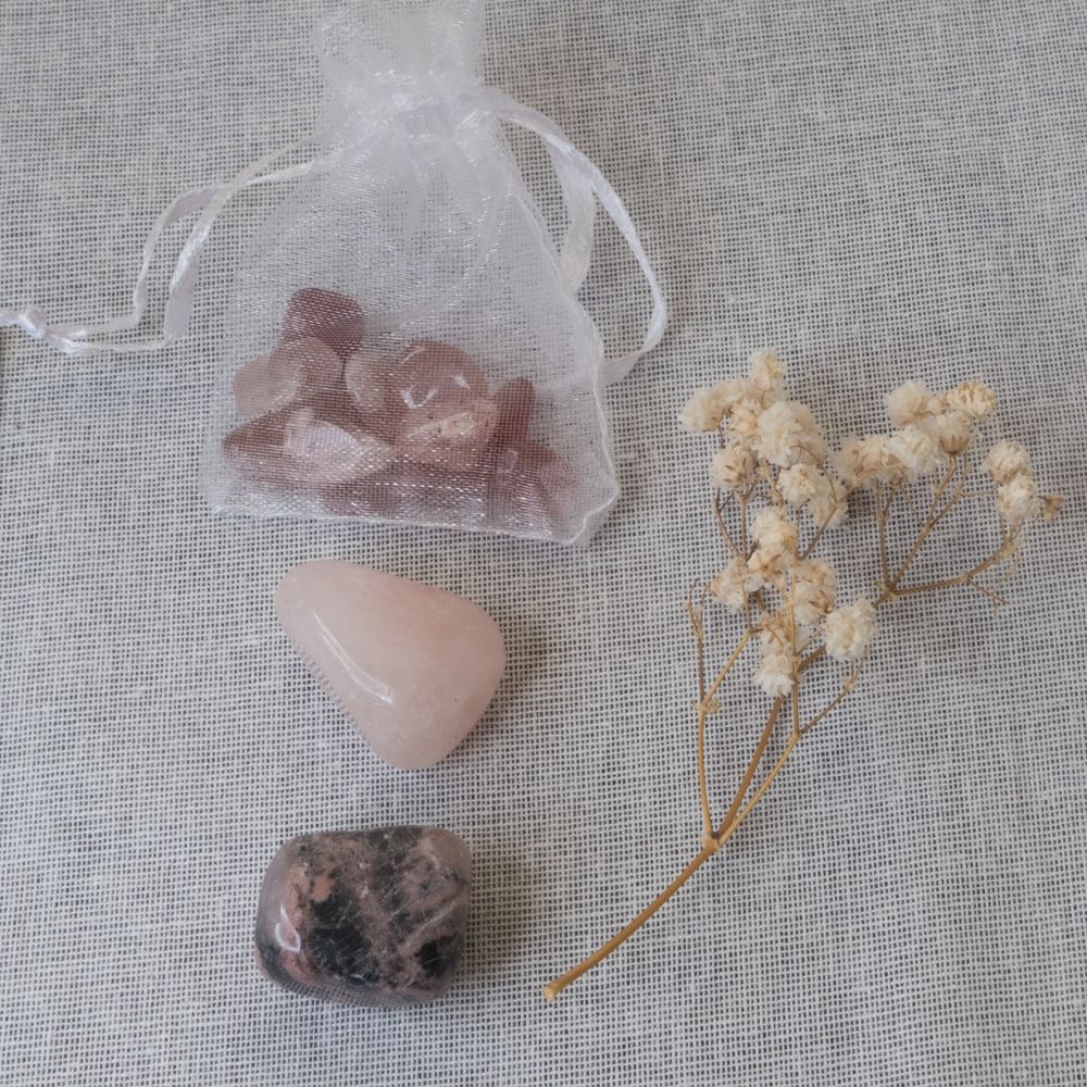 a rose quartz tumblestone, a rhodonite tumble stone and some strawberry chips , a branch of dried flowers on a textured fabric background