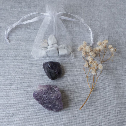 howlite chips, amethyst tumble stone and lepidolite rough stone on a white surface with a clear bag and dried flowers.