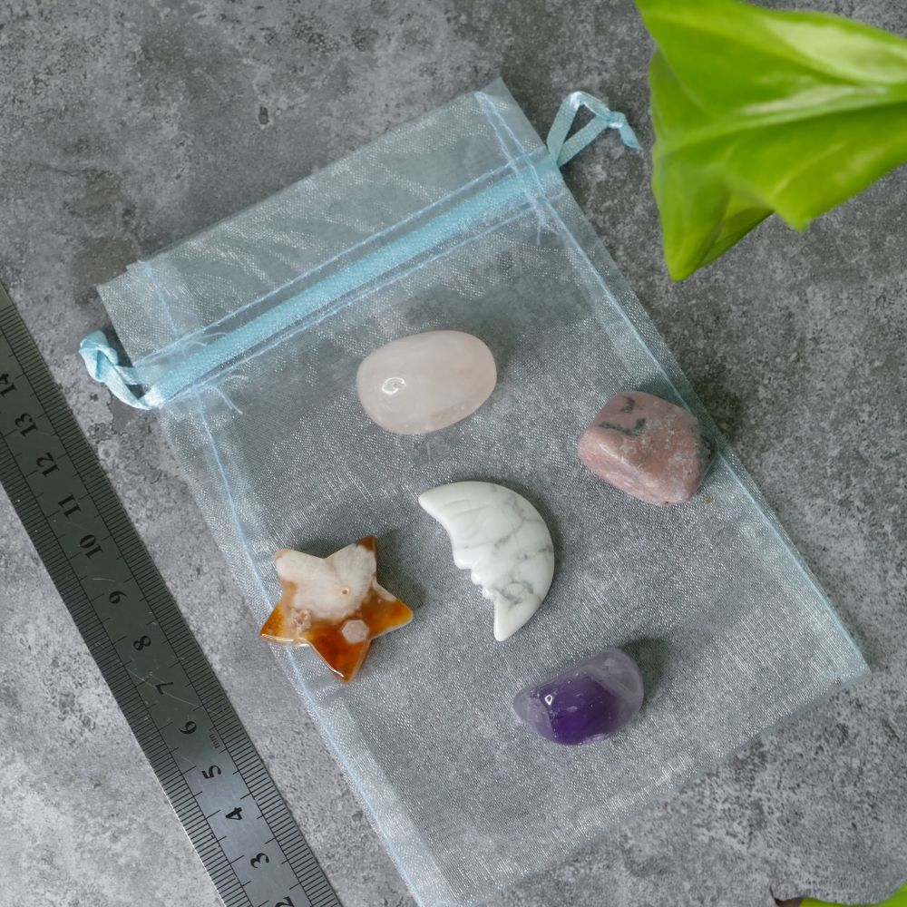 Clear drawstring bag with rose quartz, rhodonite, amethyst tumbled stones, a howlite moon and a flower agate star on a gray surface with a ruler for scale.