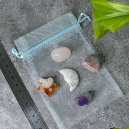 Clear drawstring bag with rose quartz, rhodonite, amethyst tumbled stones, a howlite moon and a flower agate star on a gray surface with a ruler for scale.