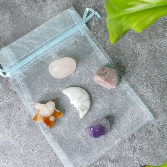 Clear drawstring bag with rose quartz, rhodonite, amethyst tumbled stones, a howlite moon and a flower agate star on a gray surface