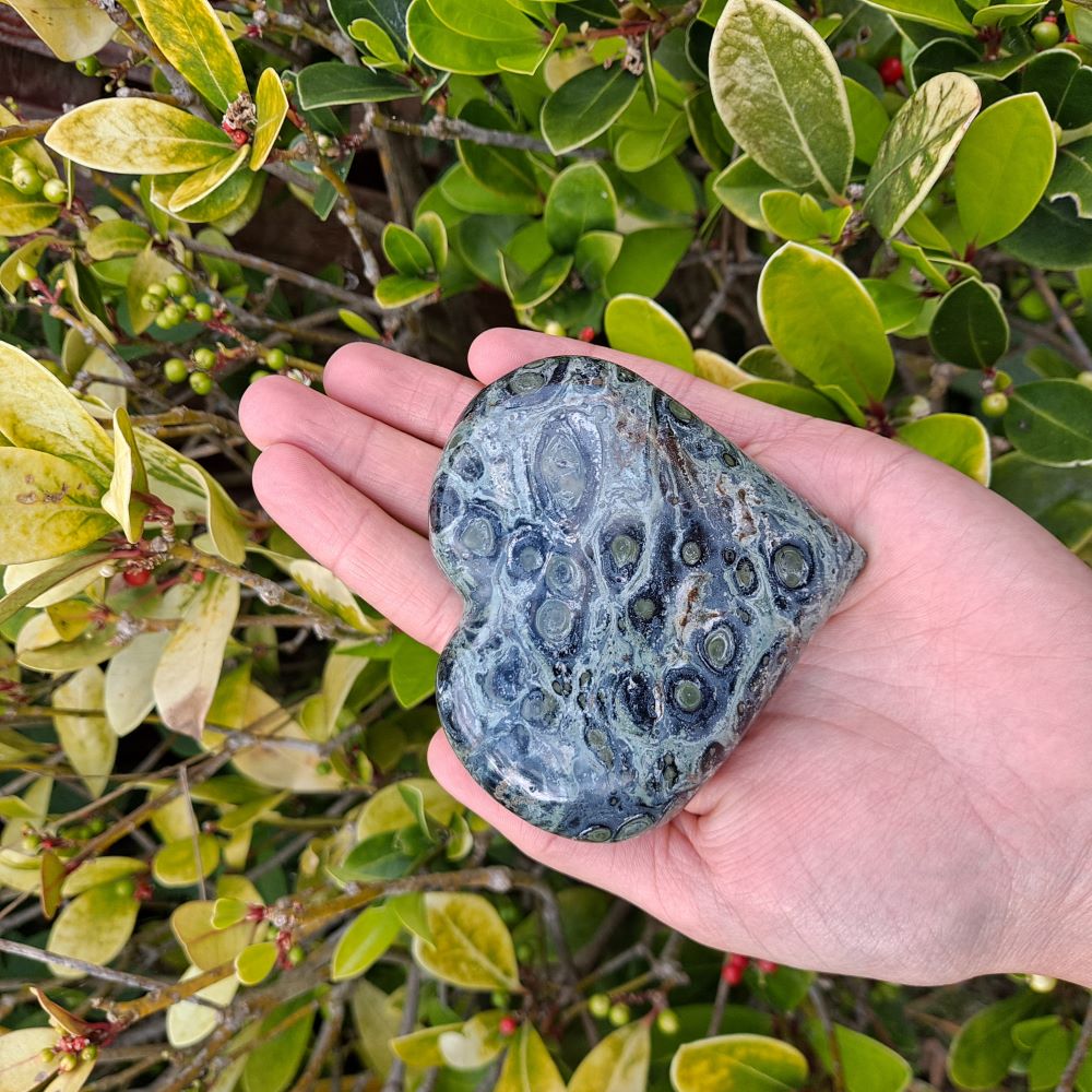 Dumi's Crystals | Unique Kambaba Jasper Heart Carving (Growth & Harmony): A stunning view of a unique Kambaba Jasper Heart Carving, a symbol of growth & harmony. Measuring 3 inches tall, its rich green hues & orbicular patterns connect you to the earth's energy. Embrace positive change with Dumi's Crystals (ethically sourced).